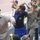 A group of young men are gathered in what appears to be a stadium or sports venue, with one central figure in a blue Inter Milan football jersey and sunglasses dancing or celebrating energetically with his arms raised. Several onlookers surround him, some smiling and participating in the festive atmosphere. A brass instrument (trumpet or horn) is visible at the bottom of the frame.