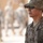 A U.S. Army soldier in the foreground is wearing Army Combat Uniform (ACU) digital camouflage, a boonie hat, and tactical sunglasses. The soldier has visible rank insignia (Staff Sergeant chevrons) and an Airborne tab on the left shoulder. Several other uniformed soldiers are visible in the blurred background, suggesting a military formation or outdoor assembly area.