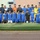 A large group of soccer/football players and officials pose for a team photo on a grass field inside a stadium. The group includes players wearing blue and yellow Adidas jerseys, a goalkeeper in orange, and several individuals in police or security uniforms. A soccer ball is visible on the ground in front of the group.