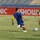 Two soccer players are competing for the ball on a grass field during a match. One player wears a blue and yellow kit while the other wears a yellow jersey with black shorts and green socks. The match is taking place in a stadium with blue seating visible in the background and a red advertising board reading 'SHOWNIC'.
