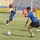 A football (soccer) match is being played on a grass pitch inside a stadium. Players in blue and yellow kits are competing, with one player wearing number 9 on their blue jersey. The stadium appears to be in the Middle East, evidenced by Arabic text on the perimeter advertising boards.