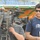 Two men are positioned near a professional broadcast camera setup at what appears to be a sporting or outdoor event. One man wearing a 'Tsunami Sportswear' t-shirt and sunglasses stands to the right, displaying a press/media credential badge. The other man, partially obscured by the camera, appears to be operating or adjusting the Sony DVCAM professional video camera mounted on a tripod.