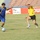 A soccer match is taking place on a grass field inside a stadium. Two opposing players are competing for the ball, with one player in a blue and yellow Adidas kit dribbling while a player in an all-yellow and black kit attempts to challenge. A third player in the same blue and yellow uniform is visible in the background.