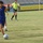 A soccer match is being played on a grass field, likely in Iraq based on the Arabic banner referencing the Iraqi Olympic Committee. A player in a blue and yellow Adidas kit is dribbling a soccer ball, while a referee in a green shirt and black shorts follows the play. Additional players and spectators are visible in the background.