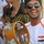 A young man is smiling broadly while holding a tambourine with metal jingles. He is wearing a white jersey with an Iraqi flag patch on the chest, dark sunglasses, and a black head wrap, with a yellow cloth tied around his wrist. The setting appears to be an outdoor public gathering or celebration.