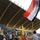 A celebratory scene at a stadium where a crowd of spectators watches from the stands while individuals on the ground level wave an Iraqi flag and a yellow flag. A person in red athletic wear and a yellow headband appears to be celebrating or dancing near the barrier wall. Military or security personnel in camouflage uniforms are present alongside civilians.