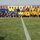 A large group of approximately 40 men pose for a team photo on a grass soccer/football field at a stadium. The group appears to consist of two soccer teams wearing blue-and-yellow uniforms, along with referees in green and black, and several individuals in military uniforms and civilian clothing. A soccer ball is visible on the center line of the field.