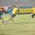 A football (soccer) match is being played on a grass pitch inside a stadium. One player in a blue and yellow Adidas kit is competing against multiple players wearing yellow jerseys and dark shorts. The background shows stadium seating, advertising banners including 'ZAIN' and Arabic text referencing the Iraqi Olympic Committee.