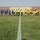 A football (soccer) match pre-game or post-game ceremony is taking place on a grass pitch, with two teams walking across the center line of the field. One team is wearing blue and yellow uniforms while the other wears yellow and black. Three referees in green/yellow and black attire are visible in the center, with one carrying a football.