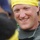 A smiling adult male wearing a yellow swim cap and what appears to be a wetsuit or athletic gear with harness straps. He appears to be at an outdoor athletic or competitive event, surrounded by other people in the blurred background. His expression is cheerful and relaxed.