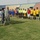 A pre-match ceremony at a stadium where two soccer teams are lined up on the field alongside referees. Military personnel in US Army combat uniforms and local security forces in camouflage uniforms are walking along the line of players, appearing to greet or inspect them. The setting appears to be a mid-sized stadium with colored seating visible in the background.