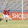 A goalkeeper in a red and navy jersey with white shorts is captured mid-action near the goal, appearing to kick or clear a soccer ball. The match is taking place in a stadium with blue empty seats in the background. Advertising banners in Arabic and English (including 'SHOWNIC') are visible along the perimeter.