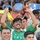 A crowd of spectators at what appears to be a sporting event, with a central figure wearing a green Iraq national team jersey celebrating enthusiastically with arms raised. Two men in Iraqi security forces camouflage uniforms are visible in the foreground. The atmosphere is festive and celebratory.