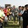 A group of U.S. military personnel in ACU camouflage uniforms are meeting with local officials and soccer players on a stadium field. A handshake or greeting exchange is taking place between a U.S. soldier and a player holding a soccer ball wearing goalkeeper gloves. The setting appears to be a formal ceremonial or goodwill event at a sports stadium.