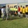 A soccer match ceremony or pre-game lineup is taking place in a large stadium. Military personnel in U.S. Army ACU camouflage and local security forces in blue digital camouflage uniforms are walking past a line of soccer players and referees. The players are wearing yellow and black uniforms, while referees are dressed in bright yellow/lime shirts.
