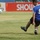 A soccer player wearing a blue jersey with the number 9 and black shorts is captured mid-kick on a grass field. The ball is visible in the air to the left, having just been struck. Advertising boards in the background display partial text including 'SHOWTIME' and 'LCD' along with a 'ZAIN' logo.