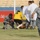 The image shows an emergency response scene on a football/soccer pitch inside a stadium. A player wearing a yellow jersey with number 4 appears to be injured on the ground, with multiple personnel including individuals in military/police camouflage uniforms and a person in a white medical coat attending to them. One security personnel in blue-grey digital camouflage is running toward the camera while others assist the downed player.