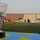 A silver trophy cup on a black base is prominently displayed in the foreground on a blue and yellow banner or flag. In the background, a football match or training session is taking place on a grass pitch inside a stadium with largely empty stands. The stadium features yellow and blue seating sections and perimeter advertising boards including a visible 'ZAIN' sponsor banner.