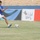 A young boy wearing a blue Adidas jersey and white shorts is running toward a soccer ball on a grass field. The scene appears to take place in a stadium with blue seating visible in the background. Advertising banners with Arabic text are displayed along the sideline.