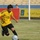 A soccer match is taking place on a grass field, with a player in a yellow jersey with black Arabic text dribbling the ball while being challenged by an opponent in a blue jersey. The stadium setting includes advertising banners in Arabic along the sideline and empty yellow seats visible in the background. The action is captured mid-play with the ball on the ground between the two players.