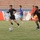 A soccer match is being played on a grass field inside a stadium with red bleacher seating visible in the background. Two teams are competing — one in black uniforms and one in blue and yellow uniforms — with a soccer ball visible mid-play. The image captures an active moment of gameplay with multiple players in motion.