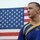 A young male with a buzz cut stands in front of an American flag being held up by another person partially visible on the left. He is wearing a blue and gold Adidas athletic jersey and appears to be speaking or reciting something. The image is a close-up portrait-style shot taken outdoors.