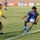 A soccer match is being played on a grass field inside a stadium. Two players in the foreground are competing for the ball, with one player in a blue and yellow Adidas kit controlling the ball near the sideline. Additional players and advertising banners are visible in the background.