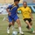 A soccer match is in progress on a grass field, with two players in the foreground competing for the ball. One player wears a blue and yellow Adidas kit, while the other wears a yellow jersey with Arabic text and black shorts. A stadium with seating and decorative murals is visible in the background.