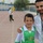A man and a young boy pose together at what appears to be an athletic stadium or sports track. The boy is wearing a green PEAK sports jersey with the number 10 and a small flag patch, while the man in a white shirt has his arm around the boy. The background shows a running track, stadium seating, and other individuals in athletic wear.
