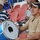 A group of men are seated in a stadium with red seats, appearing to be part of a musical support group or fan band. The central figure holds a blue bass drum branded 'AL-SADAKA Music Workshop', while another man in the background holds drumsticks near a snare drum. A third man to the left is near a brass trumpet instrument.