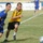 Two players in matching blue and yellow uniforms compete for position on a grass soccer field, with one player wearing black shorts labeled 'ARMY'. A soccer ball is visible in the foreground right, and a third player is visible in the blurred background. Advertising banners with Arabic text line the background of what appears to be a stadium.