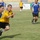 A soccer match is being played on a grass field, with players wearing blue-and-yellow Adidas jerseys competing against each other. The foreground shows two players in close pursuit, with one in a yellow jersey wearing shorts labeled 'ARMY'. A soccer ball is visible in the right portion of the frame near another player, with stadium seating and Arabic-language advertising banners visible in the background.