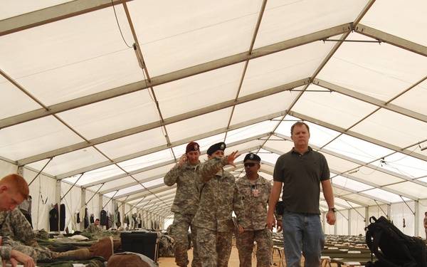 The image shows the interior of a large military tent or temporary barracks facility with rows of cots and military gear. Several soldiers in US Army combat uniforms and berets are walking through the facility alongside a civilian in civilian clothes. Additional soldiers are visible in the background, and military equipment and duffel bags are staged along the sides.