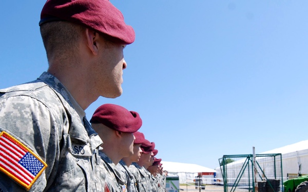 A formation of U.S. Army Airborne soldiers wearing maroon berets and Army Combat Uniforms (ACUs) stand at attention in a line. The unit patch visible on the lead soldier's left shoulder identifies them as members of the 82nd Airborne Division. A non-commissioned officer or officer stands facing the formation, apparently addressing or inspecting the troops at what appears to be a military installation or event site.
