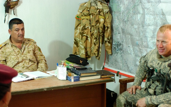 A military meeting is taking place in a sparse office or command post between personnel in different uniforms. One soldier in US Army ACU (Army Combat Uniform) with full tactical gear is seated across from personnel in desert camouflage uniforms, one of whom sits behind a desk. The setting appears to be a liaison or coordination meeting, with a map visible on the wall behind the desk.