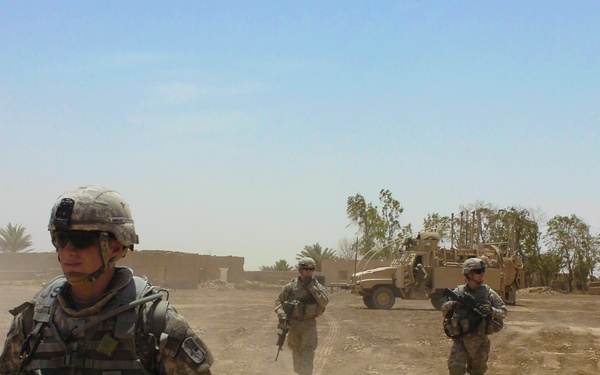 Three soldiers in U.S. Army combat uniforms conduct a dismounted patrol along a dusty dirt road in an arid environment. Military vehicles, consistent with MRAPs or HMMWVs, are staged in the background. The setting appears to be a rural area with sparse vegetation and low mud-brick structures.