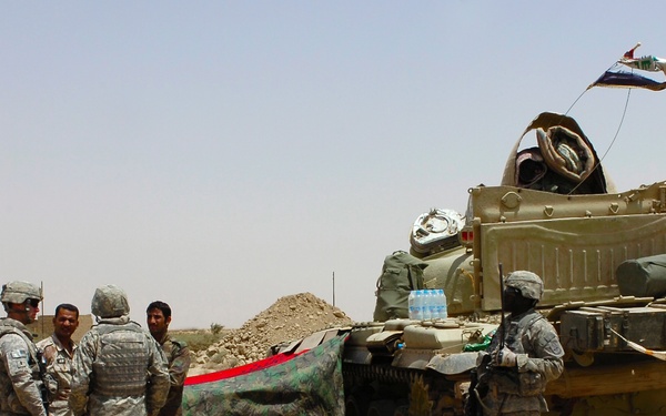 The image shows a group of military personnel gathered near a tank in an arid desert environment. Several soldiers in US Army ACU (Army Combat Uniform) camouflage are interacting with two civilians near what appears to be a Soviet-era tank flying a flag. The setting suggests a joint military operation or checkpoint interaction in a Middle Eastern desert region.
