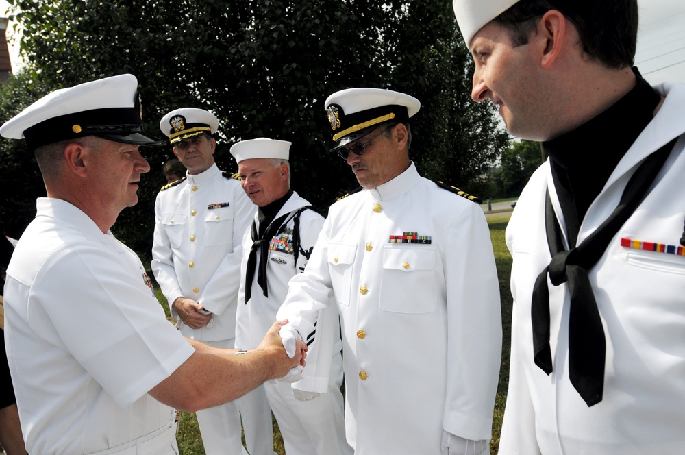 A group of United States Navy personnel in white dress uniforms are gathered outdoors in what appears to be a formal or ceremonial setting. A senior officer on the left is shaking hands with an enlisted sailor, while other officers and enlisted personnel observe. The scene suggests a formal military ceremony, inspection, or reception event.