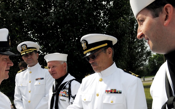 A group of United States Navy personnel in white dress uniforms are gathered outdoors in what appears to be a formal or ceremonial setting. A senior officer on the left is shaking hands with an enlisted sailor, while other officers and enlisted personnel observe. The scene suggests a formal military ceremony, inspection, or reception event.