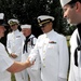A group of United States Navy personnel in white dress uniforms are gathered outdoors in what appears to be a formal or ceremonial setting. A senior officer on the left is shaking hands with an enlisted sailor, while other officers and enlisted personnel observe. The scene suggests a formal military ceremony, inspection, or reception event.