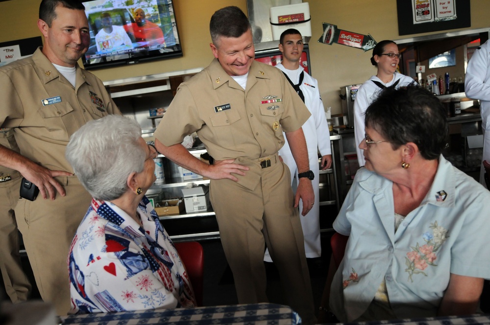 A group of U.S. Navy personnel in tan service uniforms are visiting a casual dining restaurant, interacting warmly with two civilian women seated at a table. A senior officer in the center is smiling and engaging in conversation with the seated guests. Additional Navy personnel in white uniforms are visible in the background near a service counter.