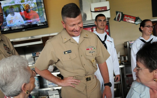 A group of U.S. Navy personnel in tan service uniforms are visiting a casual dining restaurant, interacting warmly with two civilian women seated at a table. A senior officer in the center is smiling and engaging in conversation with the seated guests. Additional Navy personnel in white uniforms are visible in the background near a service counter.
