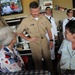 A group of U.S. Navy personnel in tan service uniforms are visiting a casual dining restaurant, interacting warmly with two civilian women seated at a table. A senior officer in the center is smiling and engaging in conversation with the seated guests. Additional Navy personnel in white uniforms are visible in the background near a service counter.