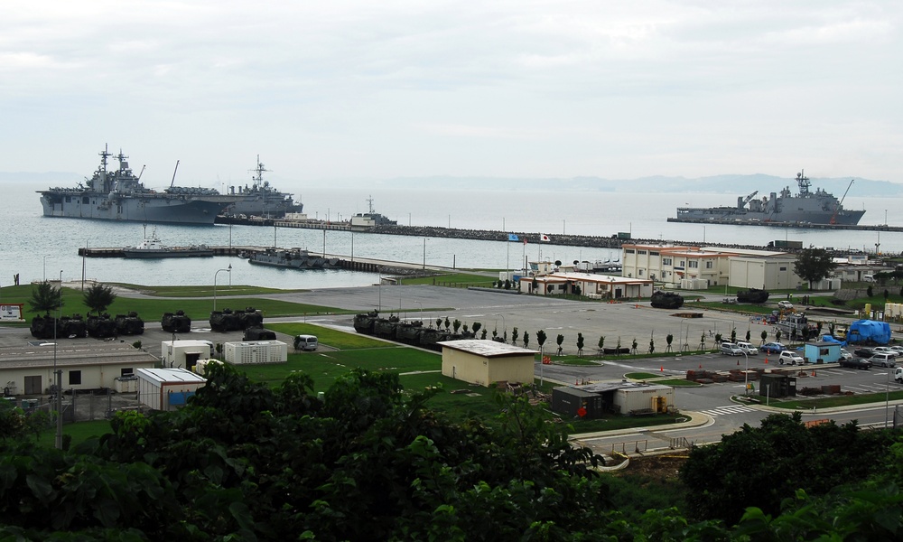 A military naval base is shown from an elevated vantage point, with multiple large amphibious assault ships moored at a pier extending into a calm bay. The shore facility includes military vehicles, administrative buildings, and paved areas consistent with a U.S. or allied naval installation. A South Korean flag is faintly visible near the pier, suggesting a joint or allied military port facility.