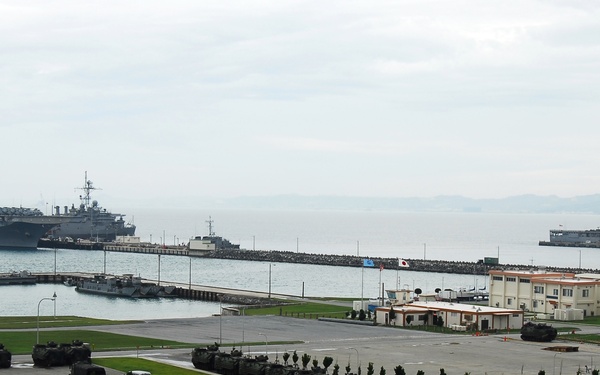 A military naval base is shown from an elevated vantage point, with multiple large amphibious assault ships moored at a pier extending into a calm bay. The shore facility includes military vehicles, administrative buildings, and paved areas consistent with a U.S. or allied naval installation. A South Korean flag is faintly visible near the pier, suggesting a joint or allied military port facility.