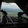 Two personnel in dark military uniforms are positioned at the open rear cargo ramp of a military transport aircraft in flight. They appear to be looking out over a landscape of forests, roads, and scattered development visible far below. The image is taken from inside the aircraft looking outward through the open ramp door.