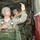 U.S. Army paratroopers are inside a military transport aircraft, preparing for a static-line parachute jump. The soldiers are in full combat gear and parachute harnesses, attaching their static lines to the anchor line cable overhead. This appears to be a standard airborne operation, likely a training or combat jump.