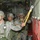 A group of U.S. Army soldiers in full combat gear and parachute harnesses are lined up inside a military transport aircraft, preparing for an airborne jump operation. The soldiers are wearing ACU (Army Combat Uniform) pattern camouflage and helmets, with static line parachute equipment visible. One soldier in the foreground is attaching or checking a yellow static line snap hook to the anchor line cable along the aircraft wall.