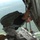 A military personnel member is positioned at the open rear ramp of a military cargo aircraft in flight, crouching near the edge while holding onto the aircraft frame. The individual is wearing military camouflage pants, a black shirt, a parachute pack, harness system, and a helmet. Green terrain is visible far below, indicating significant altitude.