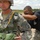 A U.S. Army airborne operation preparation scene showing soldiers being inspected before a parachute jump. A jumpmaster in a black t-shirt is conducting a pre-jump equipment check on a soldier wearing full parachute gear and ACU uniform. Multiple soldiers are visible in the background in a staging area.